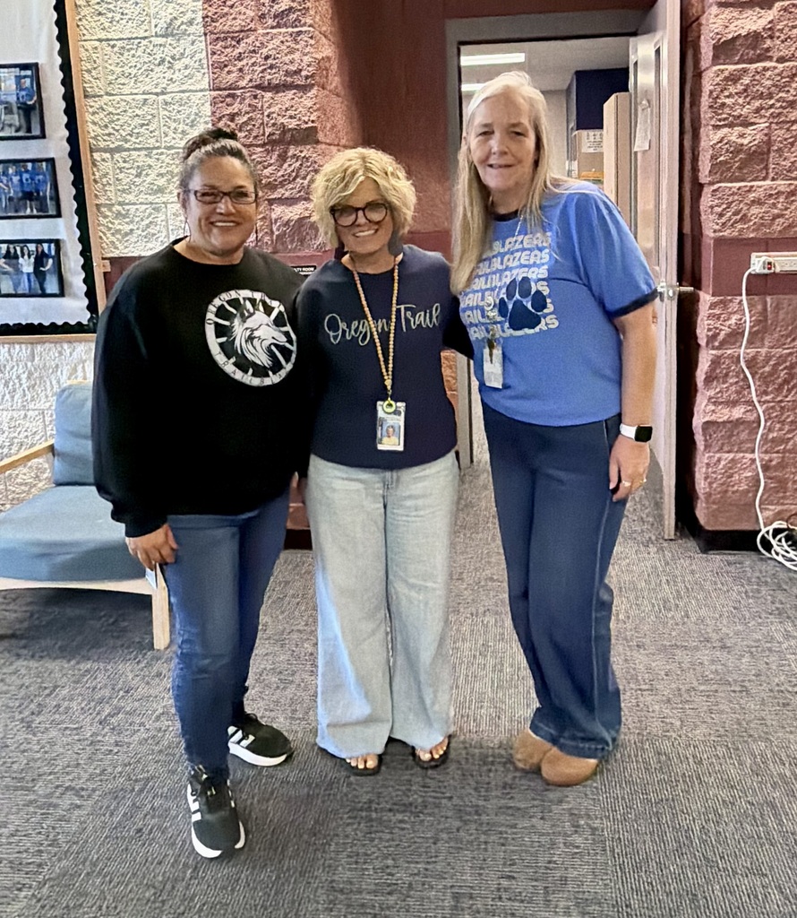 Three women in black, blue, and white shirts stand in a room with brick walls. One wears glasses and sneakers.