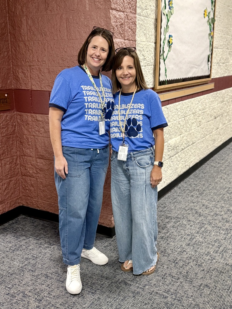 Two women in matching blue shirts and jeans stand in a hallway. One wears a watch and a necklace. A wall and picture frame are behind them.