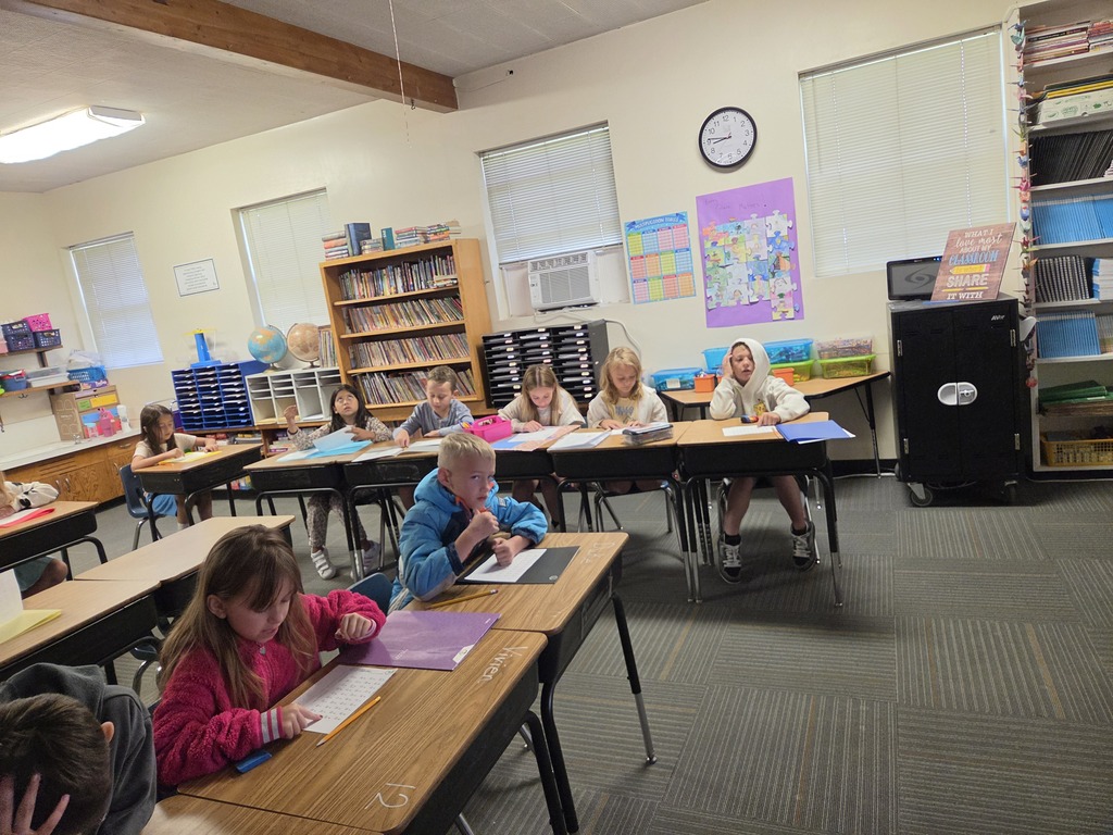 A classroom with students seated at desks. There are books and papers on the desks. A clock is on the wall.