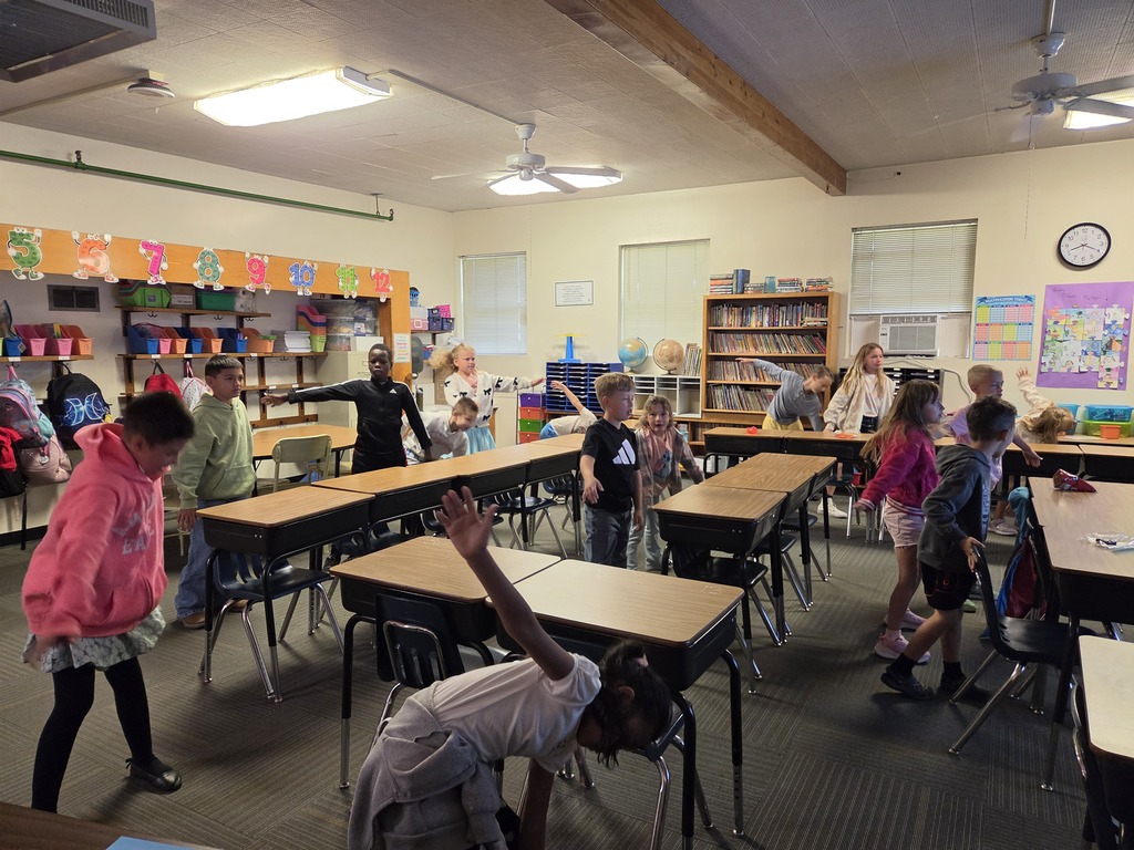 Kids participate in a class activity in a classroom. Tables and chairs are arranged, and shelves with books are visible.
