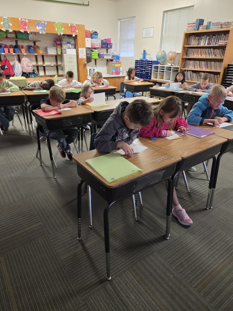 Classroom with children seated at desks, engaged in writing activities. A bookshelf with books in the background.