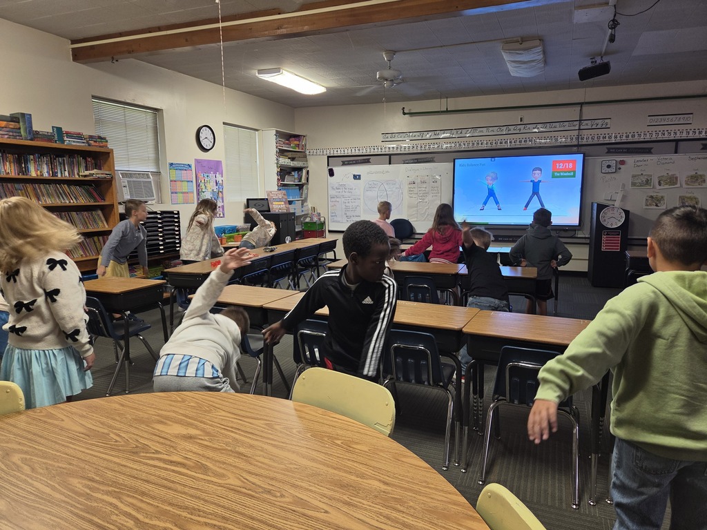 A classroom with children standing and raising their hands, watching a large screen. Tables and chairs are arranged, and a bookshelf is visible.