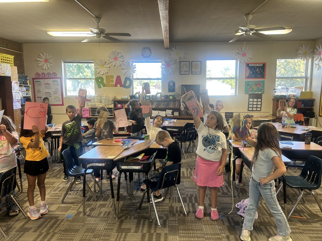 A classroom with children standing and holding up papers. Tables and chairs are arranged. Windows with bright light.