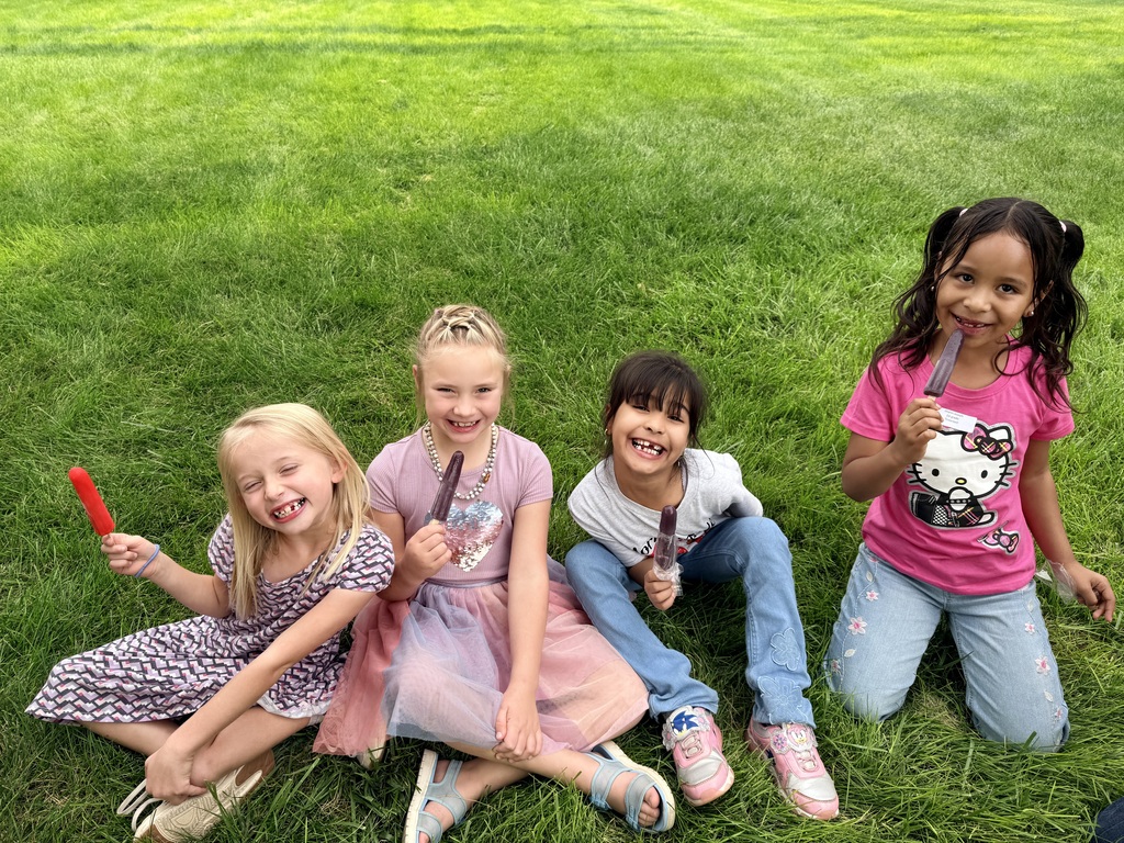 Five young girls with ice pops sit on green grass. One holds a lollipop, others have popsicles.