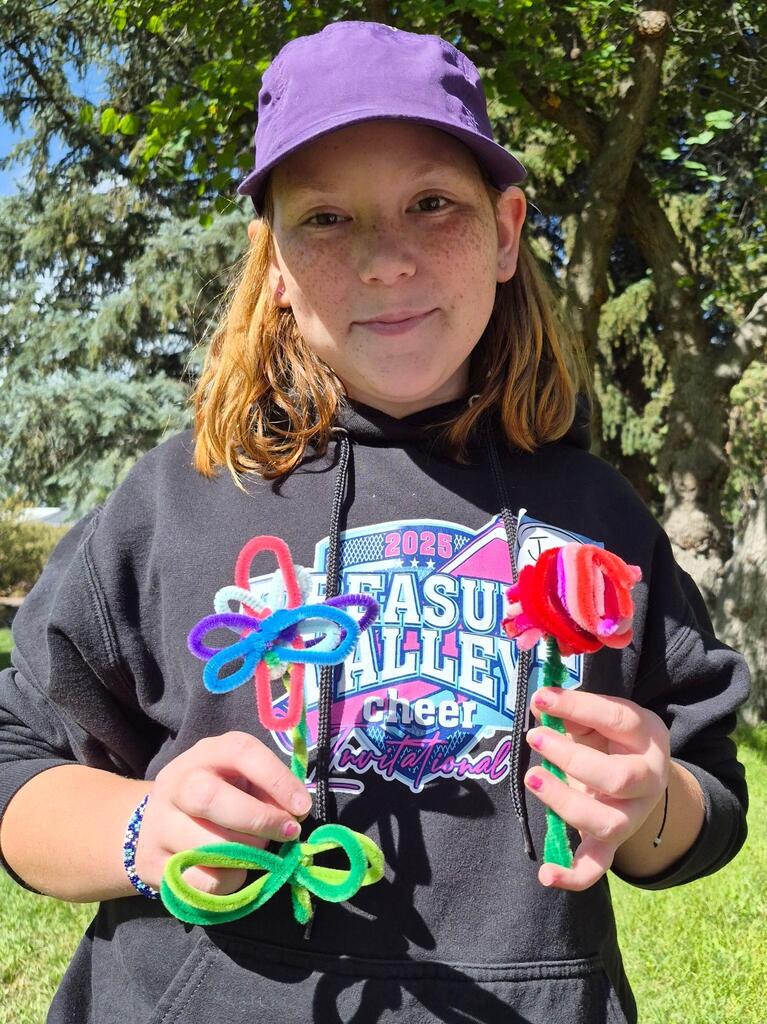 Girl in black sweatshirt and purple cap holds two colorful craft sticks shaped like flowers in front of trees.