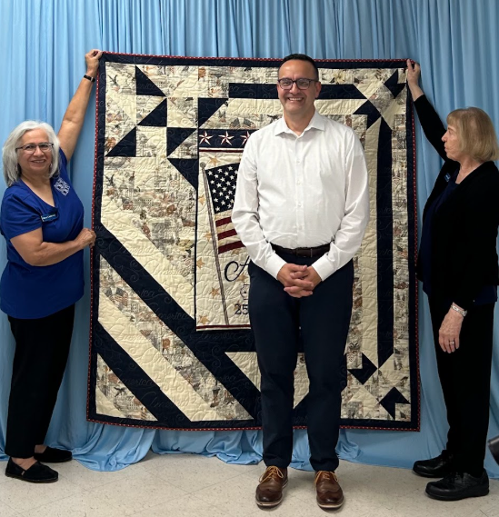Two women hold up a quilt with an American flag in the center behind Jorge Pullerio, Bickel Elementary Principal,.