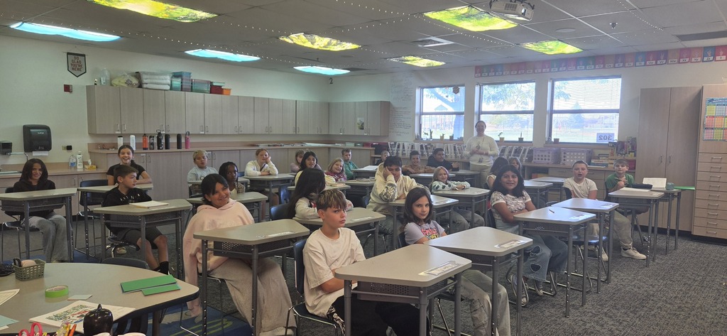 A classroom with rows of desks and students seated, facing a teacher at the front. Windows and fluorescent lights are visible.