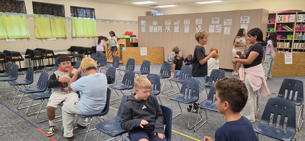 Classroom with children sitting in chairs. Some children are seated while others are standing and interacting.