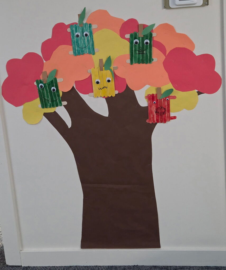 A tree-themed artwork on a white wall with a brown trunk. It has colorful leaves and several crafted apples.