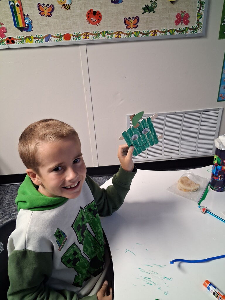 A child holding a green object with pictures of insects, sitting at a table with a cup and other items.
