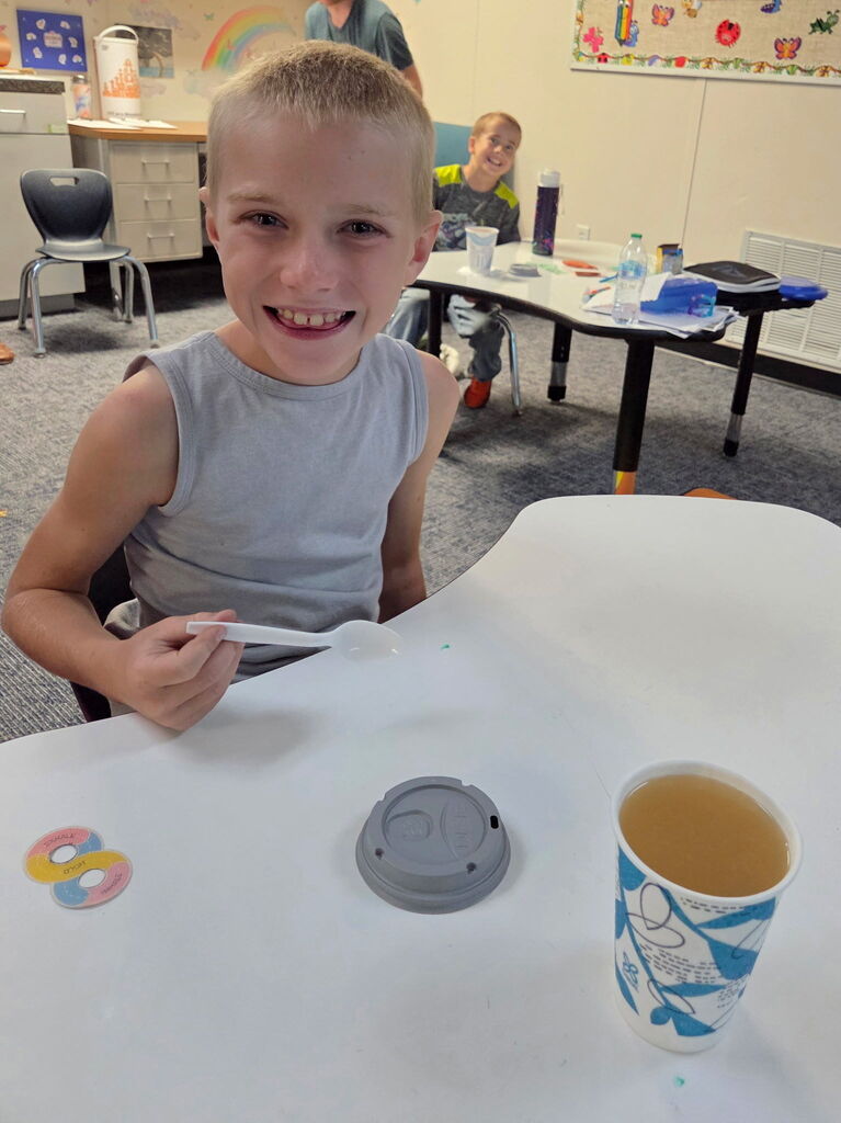 A boy smiles and sits at a table, holding a marker. A cup, a coaster, and a design are on the table.
