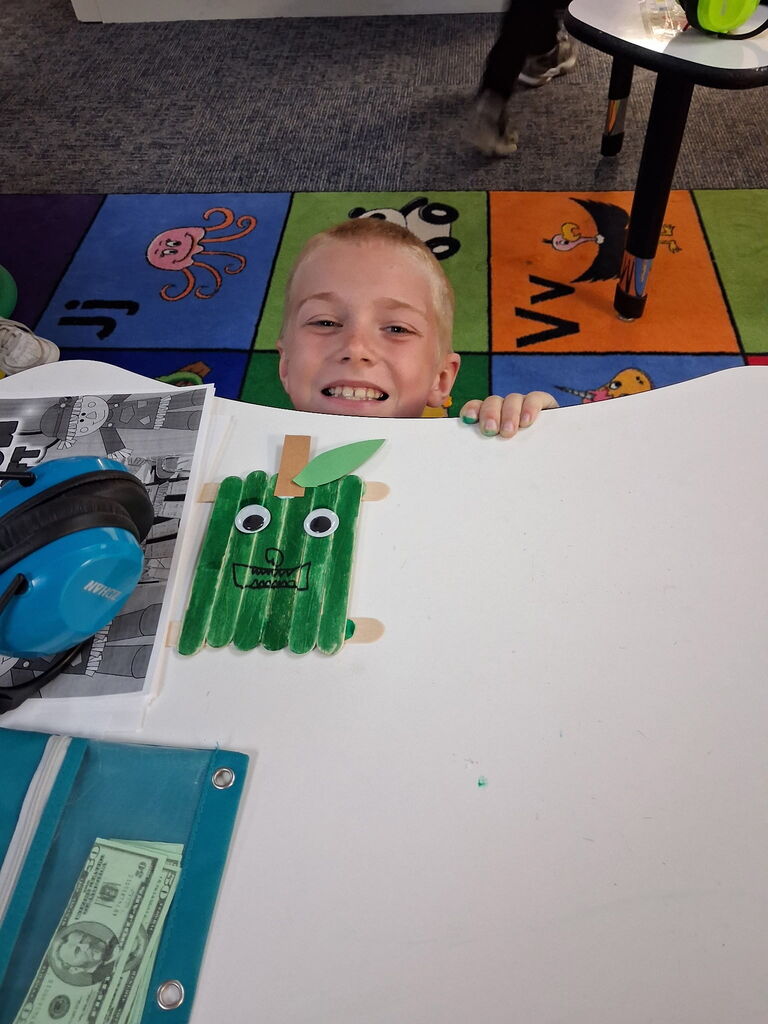 A smiling boy displays a green craft with eyes at a table with colorful rug and books.