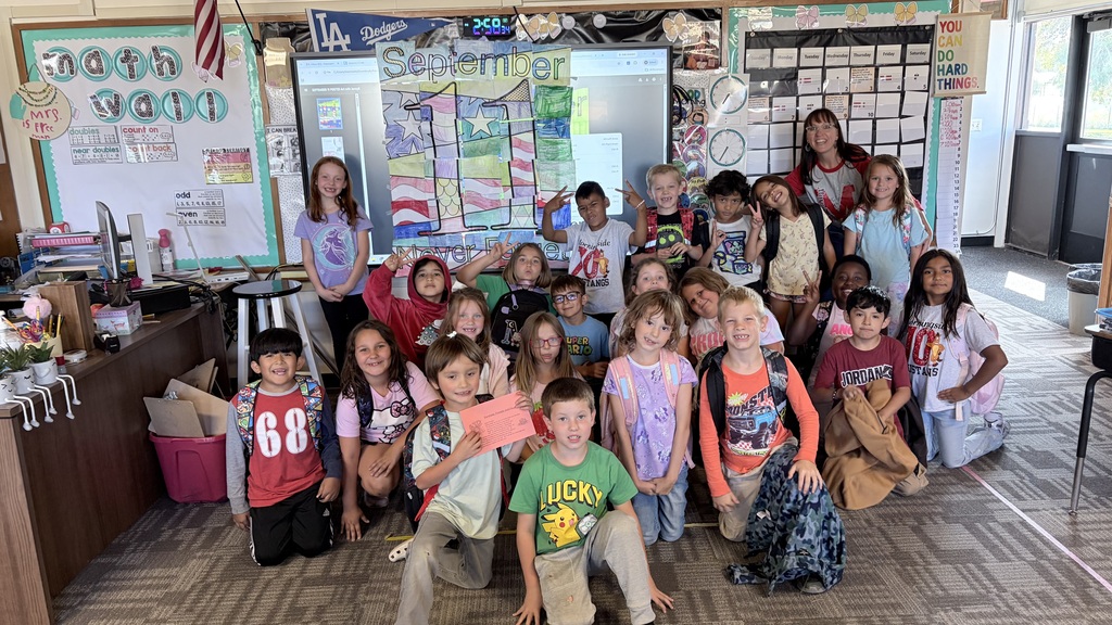Kids in a classroom wearing backpacks, holding papers, and standing and sitting around a teacher.