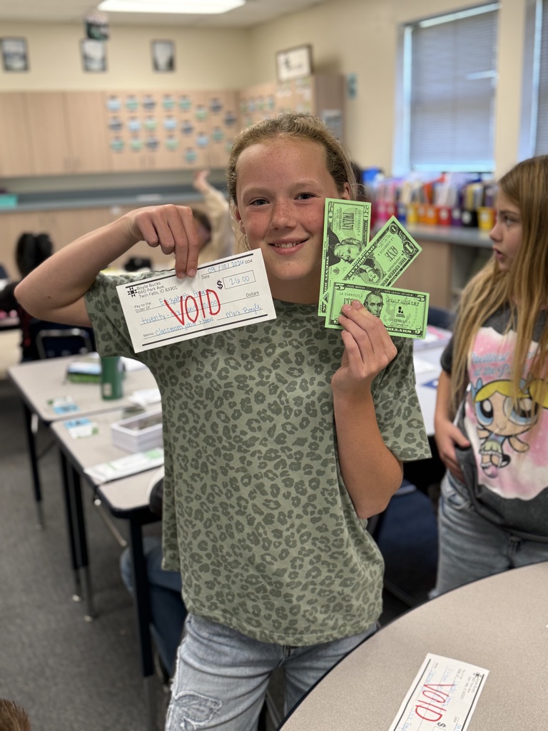 Two students in a classroom, one holding a "VOID" paper and money, smiling and standing next to a table.