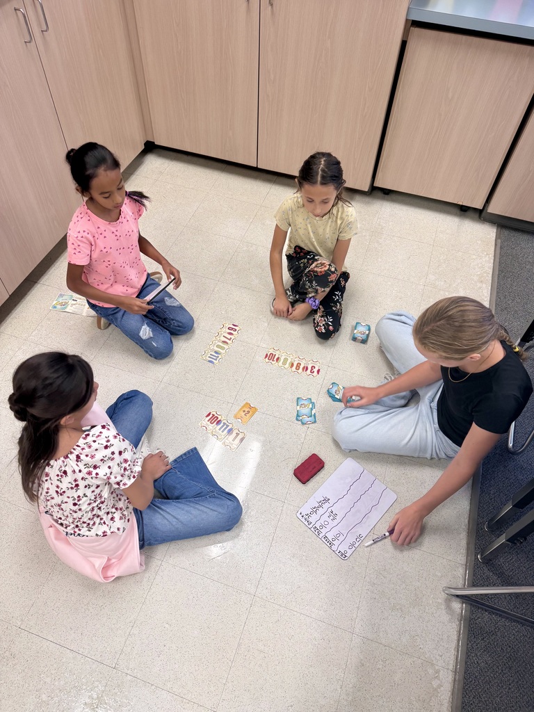 Four girls sit in a circle on a floor, with cards and a pen on the floor.