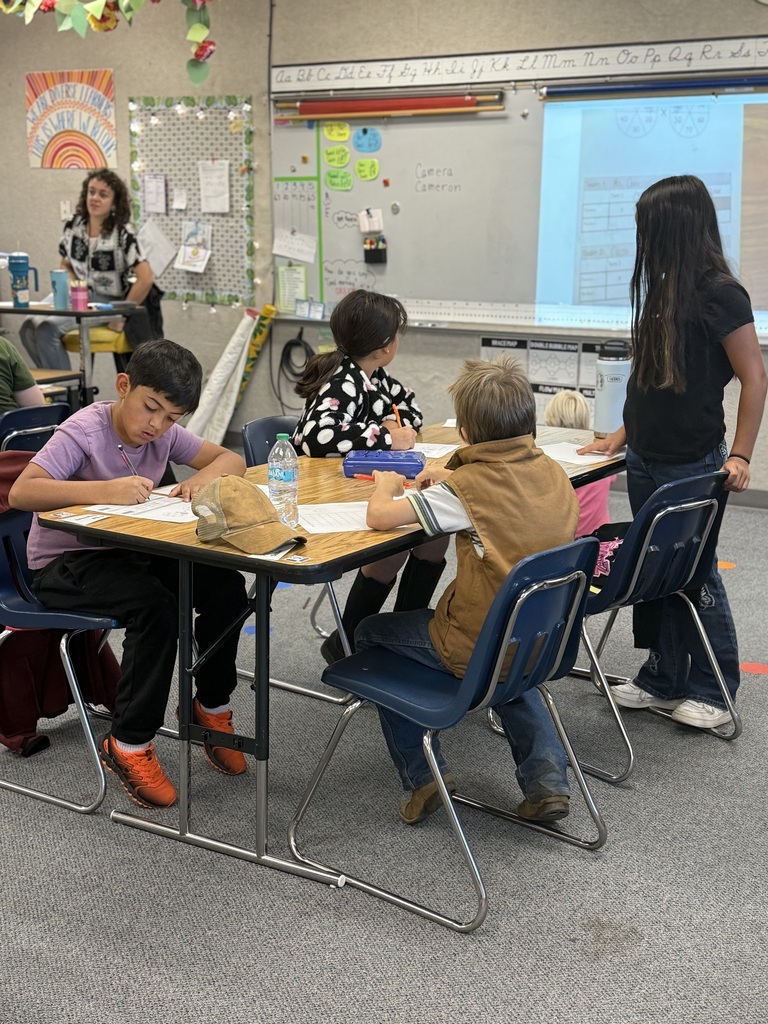 Five children, some seated at desks, work on assignments. A woman stands in the background by a whiteboard.