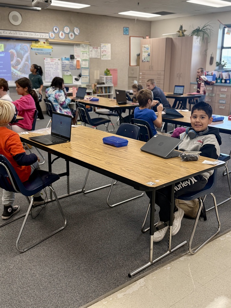 A classroom with children seated at tables using laptops. Desks have laptops, a tablet, and a mouse.