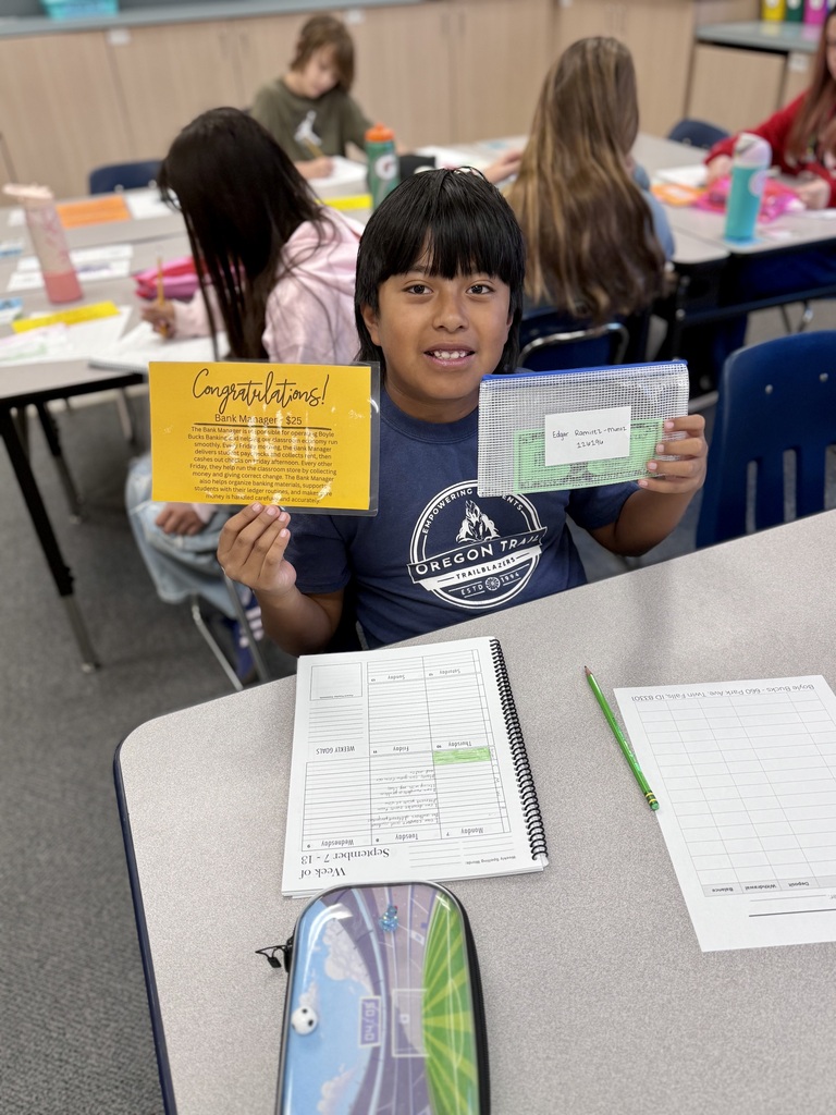 A young child sits at a table holding a certificate and a folder, surrounded by others in a classroom setting.