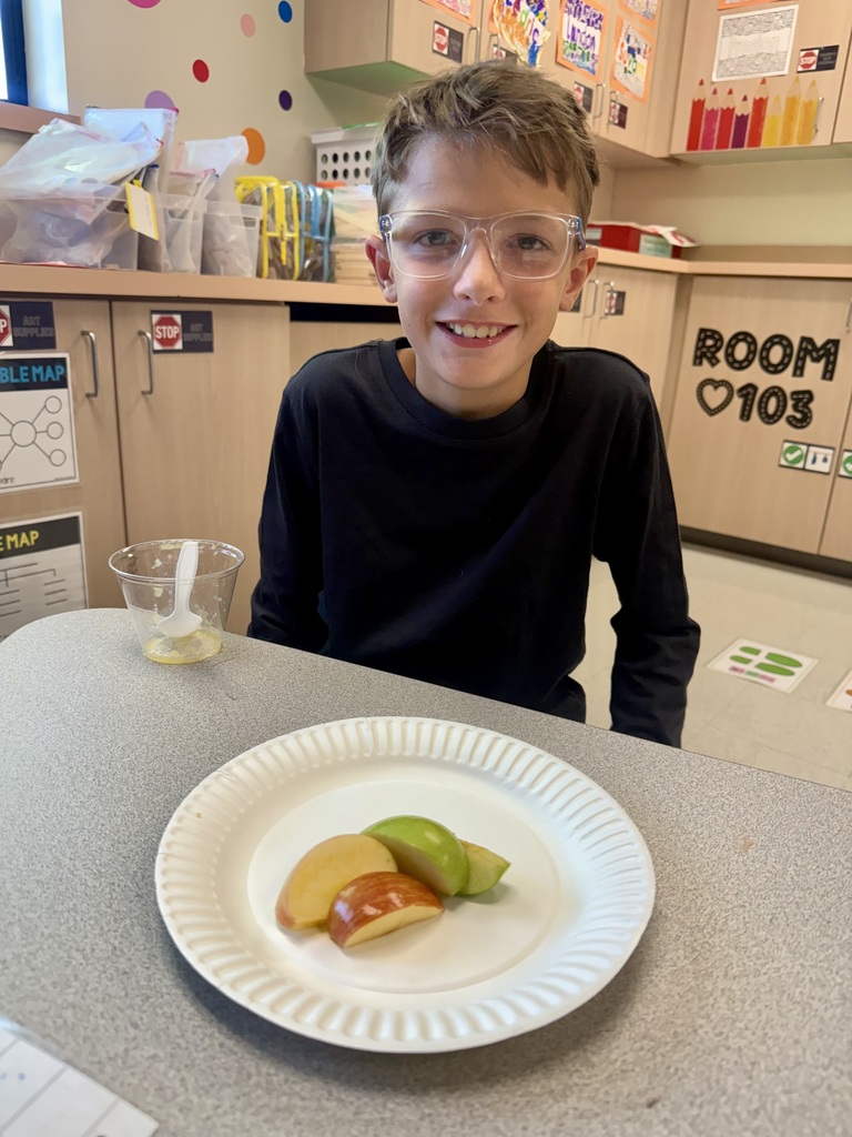 A child wearing glasses sits at a table with a paper plate containing cut apple slices.