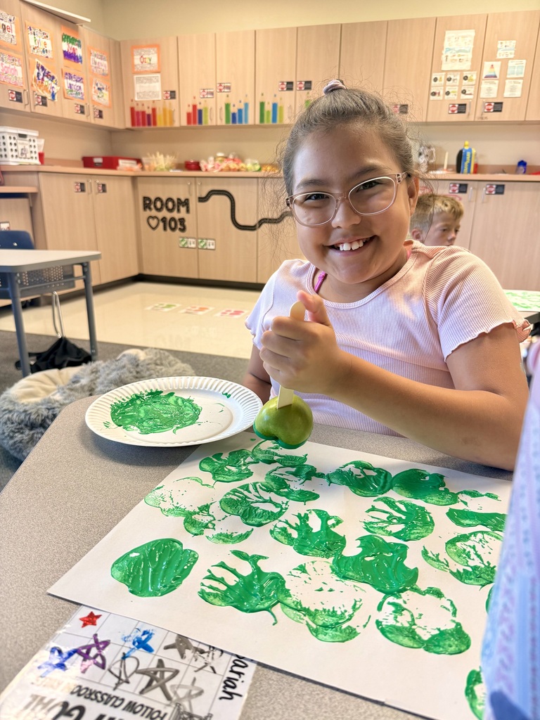A young girl wearing glasses sits at a table, painting green handprints on paper. Behind her, a classroom has shelves with various items.