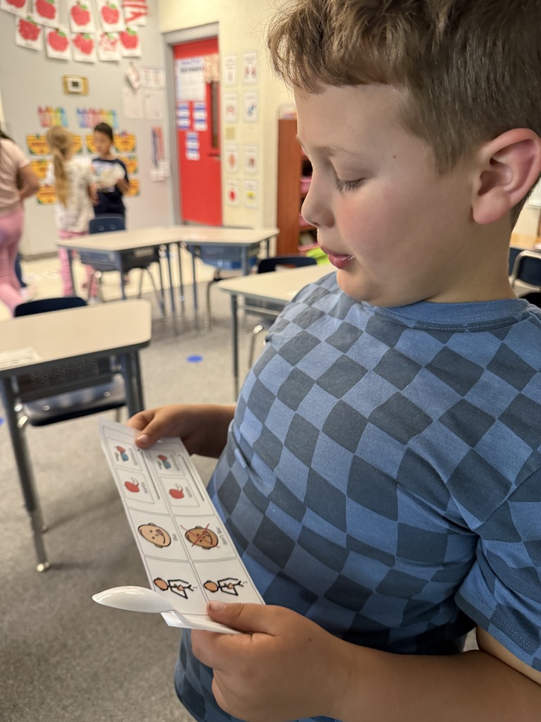 A child in a blue checkered shirt is holding a card with pictures while looking down in a classroom.