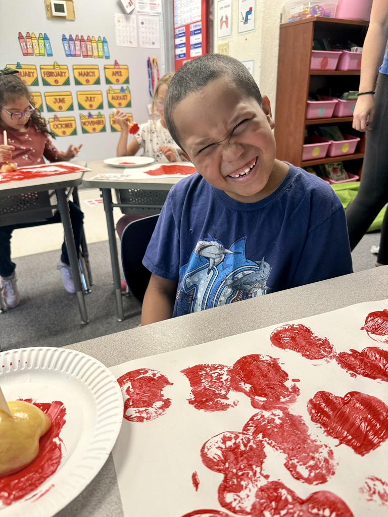A child seated at a desk, wearing a blue shirt, smiles while painting red heart shapes on paper.