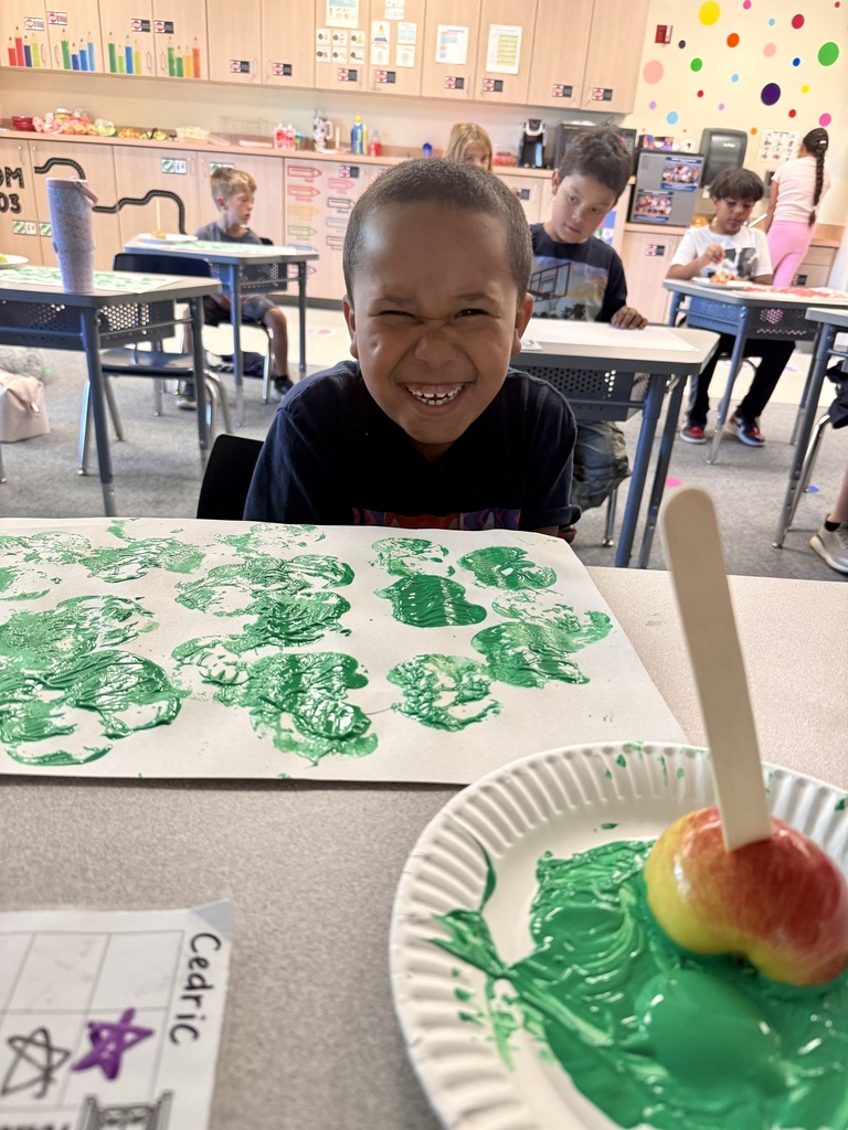 A boy sits at a desk in a classroom. In front of him is a paper with green paint and a stick.