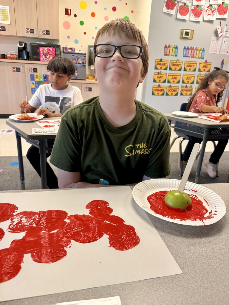A student with glasses and a green T-shirt sits at a table, facing the camera. The table displays two plates with red apple prints.