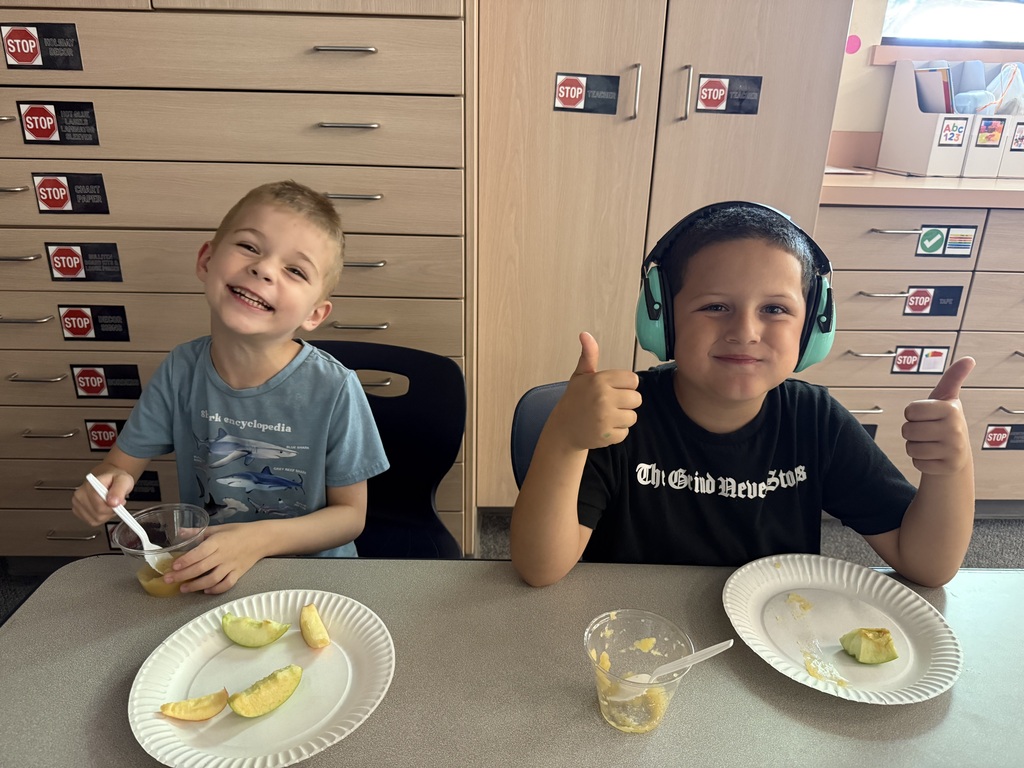 Two young boys in headphones sit at a table. One gives a thumbs up, and the other holds a spoon.