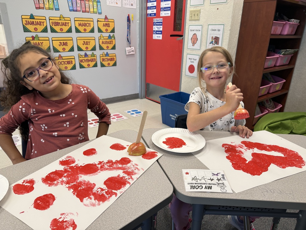 Two girls in glasses seated at a table in a classroom. They have paint on their hands and tables.