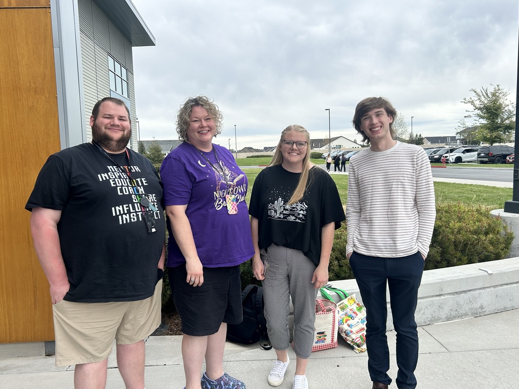 Four people stand outside a building on a sidewalk with bushes and a parking lot in the background.