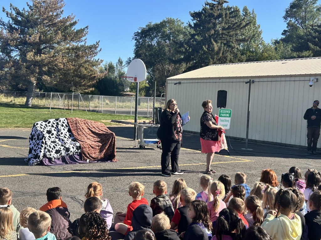 Two women speak to a crowd of children outdoors; a basketball hoop and trees are in the background.