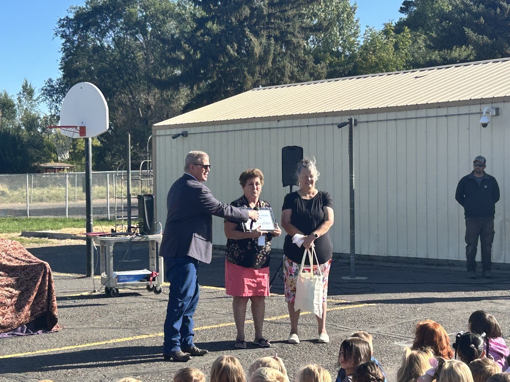 A man presents an award to two women as an audience of children watch. The award presentation occurs outdoors near a basketball court.