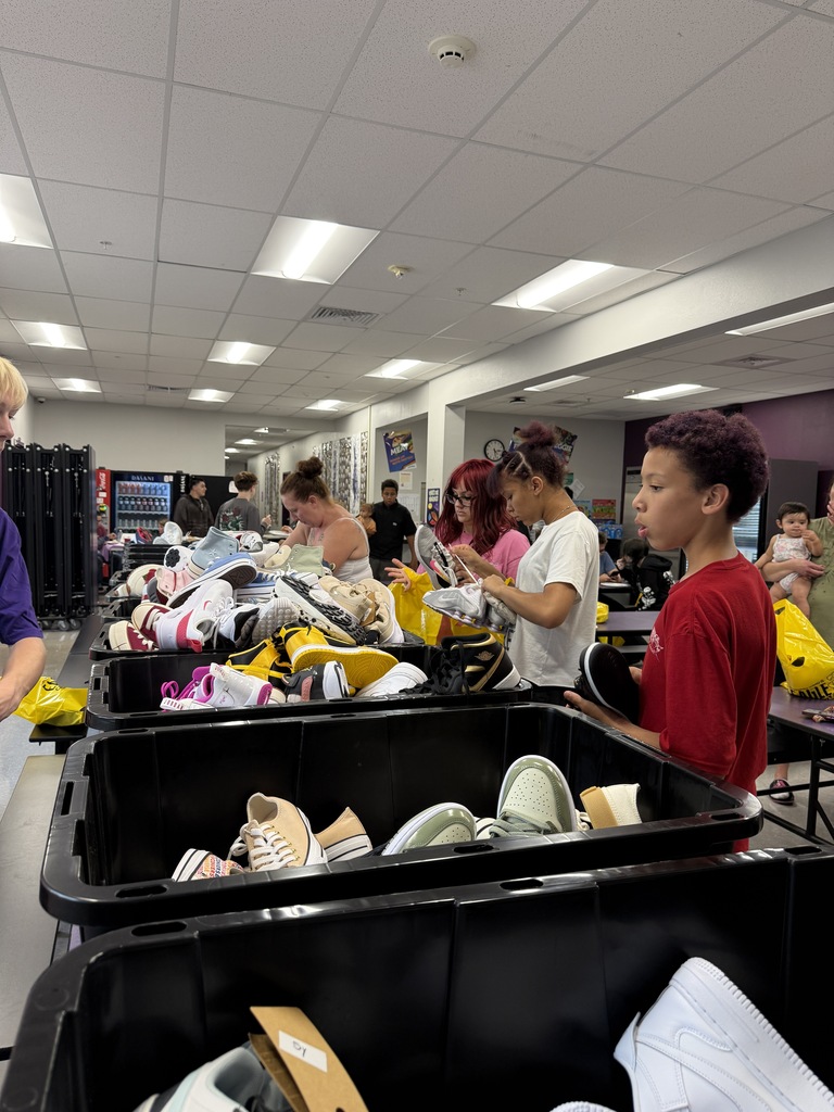 Several people stand at tables with bins of shoes and clothing. A woman in white and a boy in red.