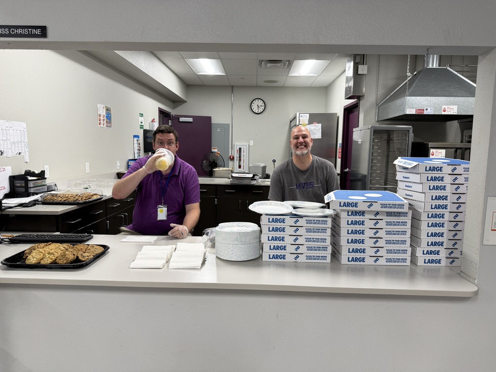 Two men in a kitchen, one sitting with a drink, other standing, with trays and plates on a counter.