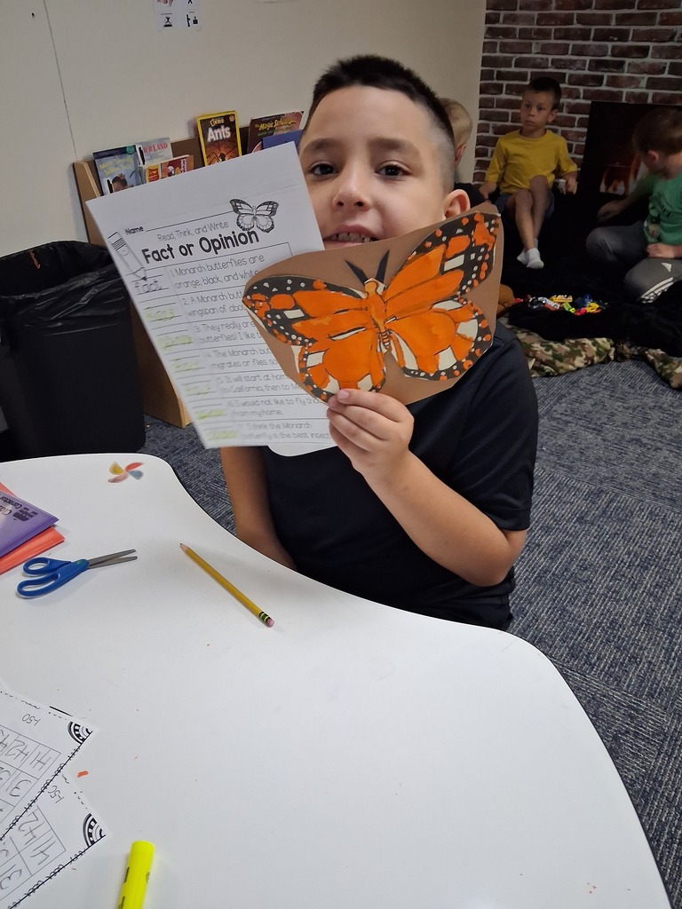 Child holding an orange butterfly cutout with wings spread out, reading a paper at a table.