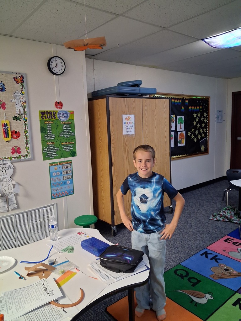A young boy in a blue shirt stands in a classroom. A white table is before him.