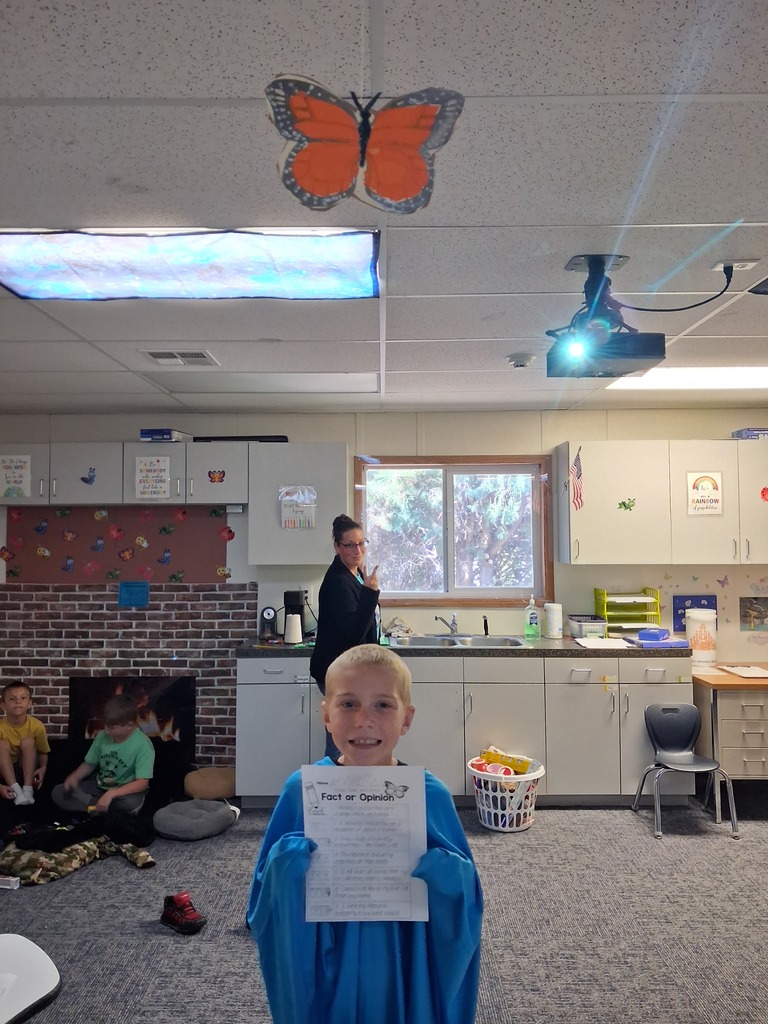 A young child in blue holds a paper in a classroom. A butterfly decoration hangs from the ceiling.