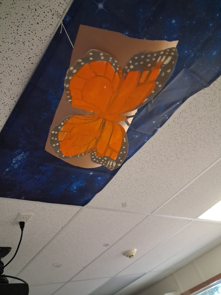 A butterfly cutout, orange with white spots, hangs on a blue backdrop with stars against a white ceiling.