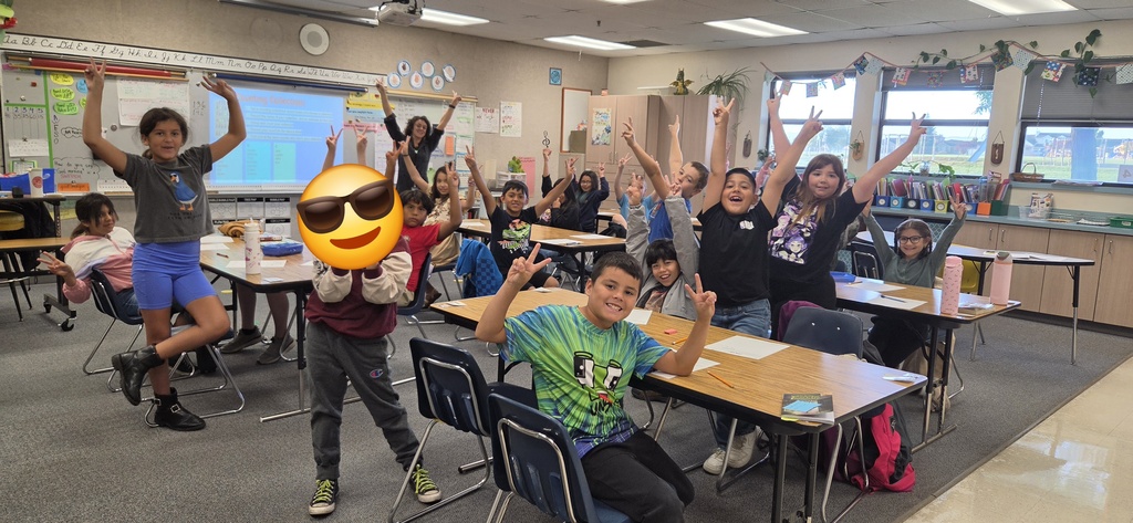 Classroom with children in casual clothes raising hands. A smiling face is superimposed on one child.