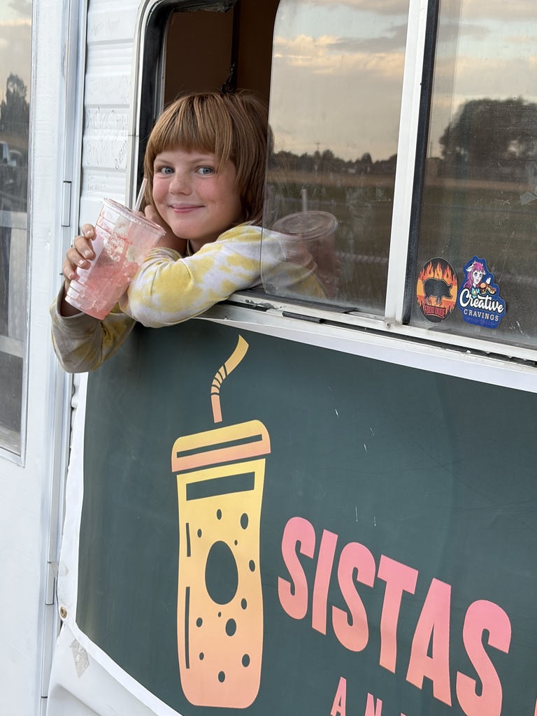 A young girl smiles while holding a drink, leaning out of a window of a food truck.