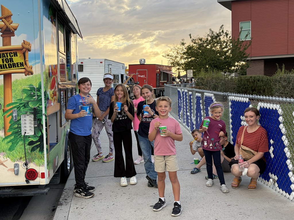 A group of people stand near a food truck, holding cups, against a fence and a sunset.