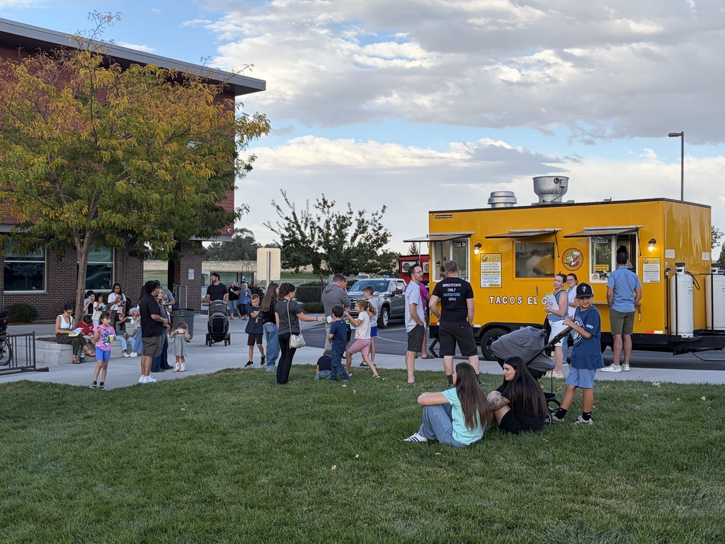 Many people gather near a yellow food truck. Some sit on grass. Trees and a building are in the background.