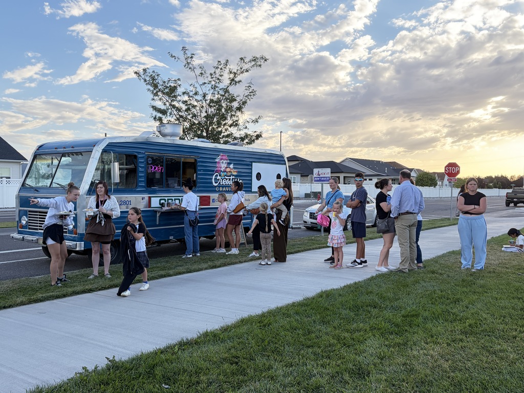 Group of people waiting in line at food truck on street with grassy area and buildings.