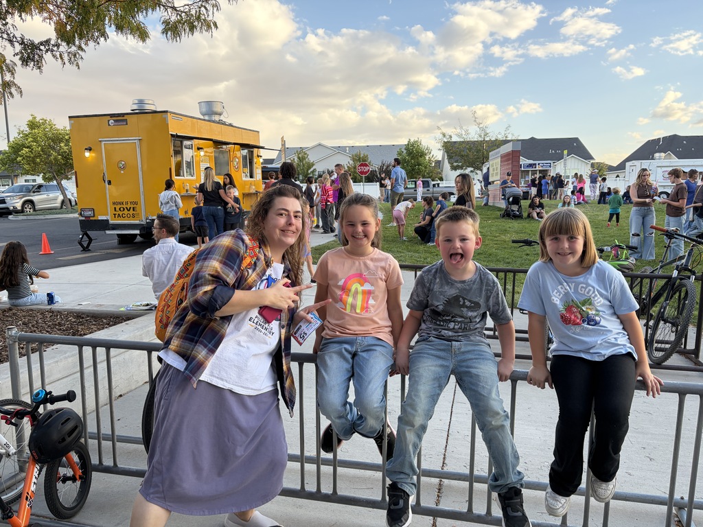 Woman and three children sit on a metal fence. A food truck and several people are in the background.