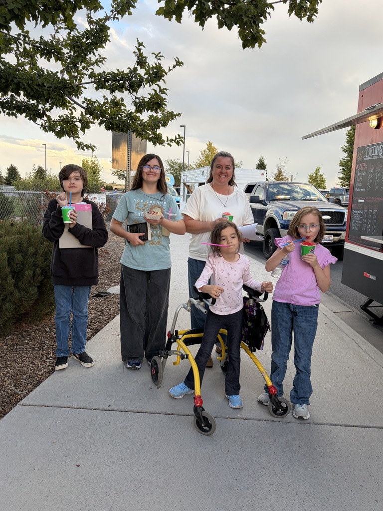 Five people stand on a sidewalk. Three children and two adults hold cups. A girl in a pink shirt uses a walker. Behind them is a food truck.