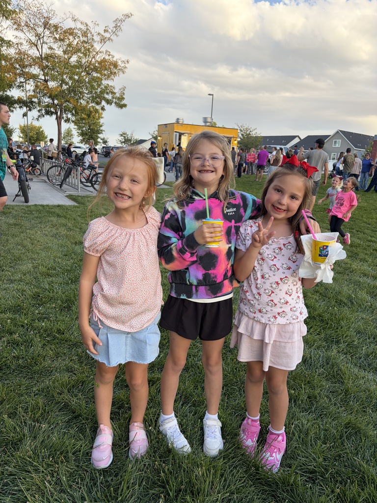 Three girls stand on grass with one holding a drink and another holding a treat. Behind them, people mill about, and a building is visible.