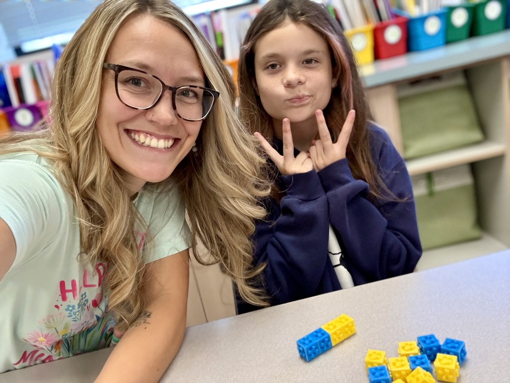 A woman and a girl sit at a table, smiling. The woman wears glasses, and the girl poses with her hands. Behind them, shelves with books and colorful bins are visible.