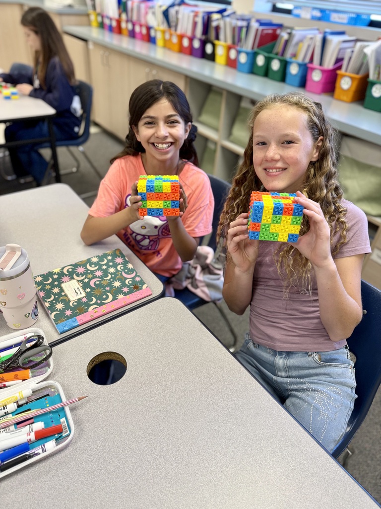 Two young girls sit at a desk holding multicolored cubes, smiling. One has a pencil box, notebook, and cup nearby. Behind them, shelves filled with colorful bins and books.