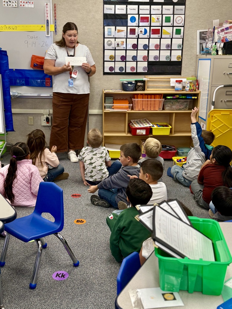 A woman reads a story to young children in a classroom, seated on the floor. There are colorful charts on the wall and shelves with various materials.